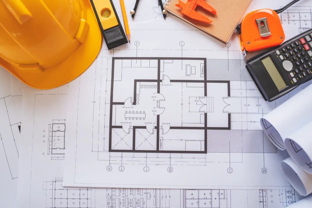 Architectural floor plan on a desk with a yellow hard hat, measuring tape, calculator, level, and drafting tools used for construction planning.