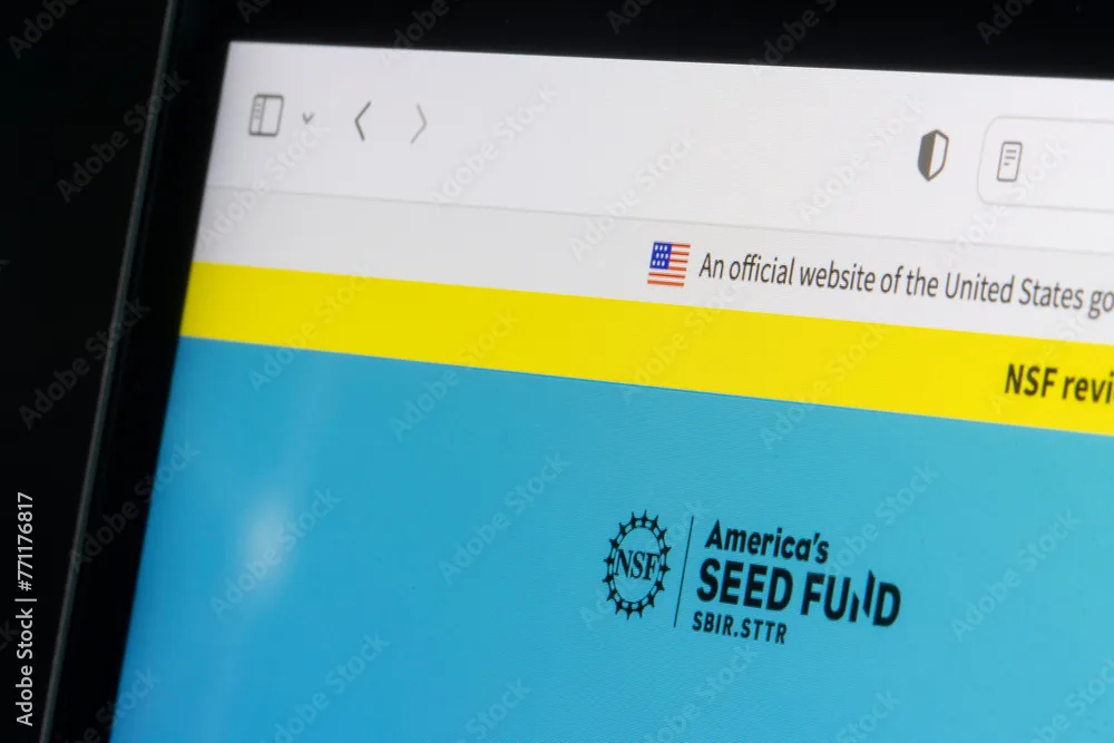 A close-up photo of a computer screen displaying a webpage from an official U.S. government site. At the top of the browser window, a small American flag icon appears next to the text “An official website of the United States government.” Below it is a bright yellow banner, and further down, part of a blue section is visible featuring the National Science Foundation (NSF) logo and the text “America’s SEED FUND SBIR/STTR.