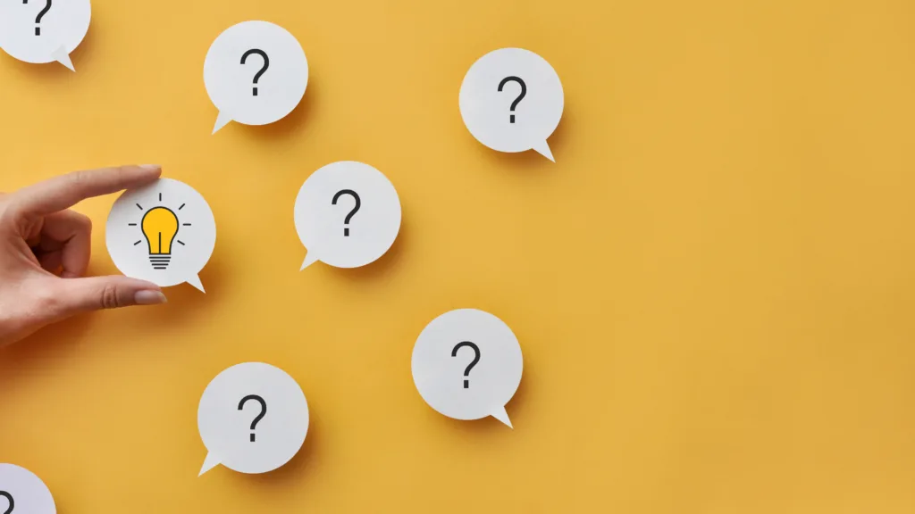 A hand holds a white speech-bubble cutout with a yellow lightbulb icon on it, surrounded by several other white speech-bubble cutouts featuring black question marks, all placed on a solid yellow background.