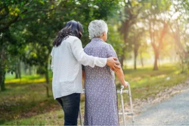 Caregiver assisting an elderly woman using a walker while walking along a park path outdoors.