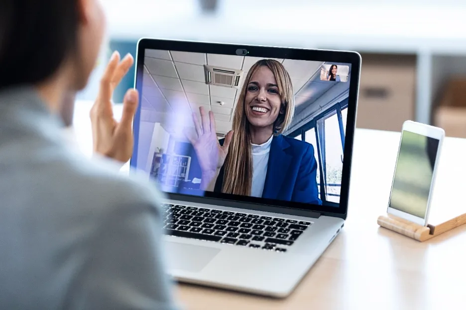 Woman participating and waving in an online video meeting from home, engaged in conversation on her laptop.
