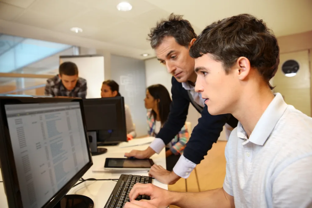 Adult man helping student on computer in classroom.