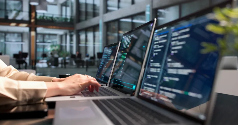 Close-up of a person typing on a laptop with lines of code displayed across multiple laptop screens in a modern office.