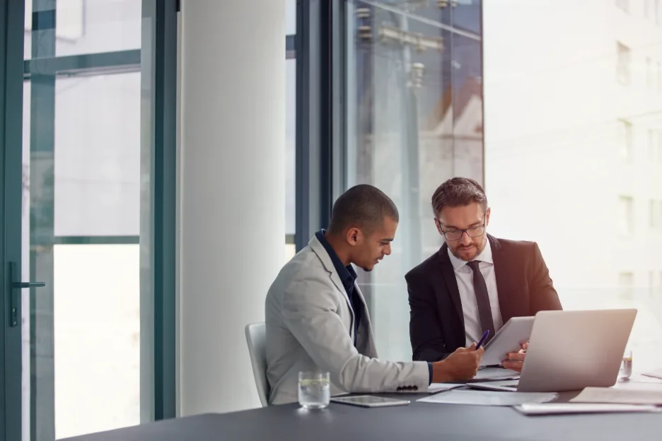 Two businessmen in suits reviewing documents and a tablet together at a conference table next to a large window.