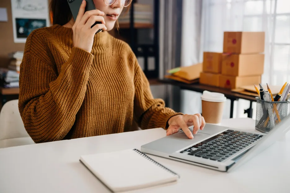 Woman talking on a smartphone while using a laptop at a desk.